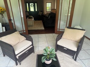 A room with two wicker chairs, a small table with a plant, and a view into a living area with a couch and a rug.