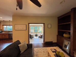 Una sala de estar con un sofá de color oscuro, un tapete blanco y mullido, y una estantería de madera. Hay una ventana con vistas a un jardín y una puerta que da a otra habitación.