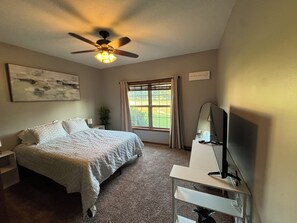 A bedroom with a bed, a ceiling fan, a window with curtains, a desk with a mirror, and a wall-mounted artwork.