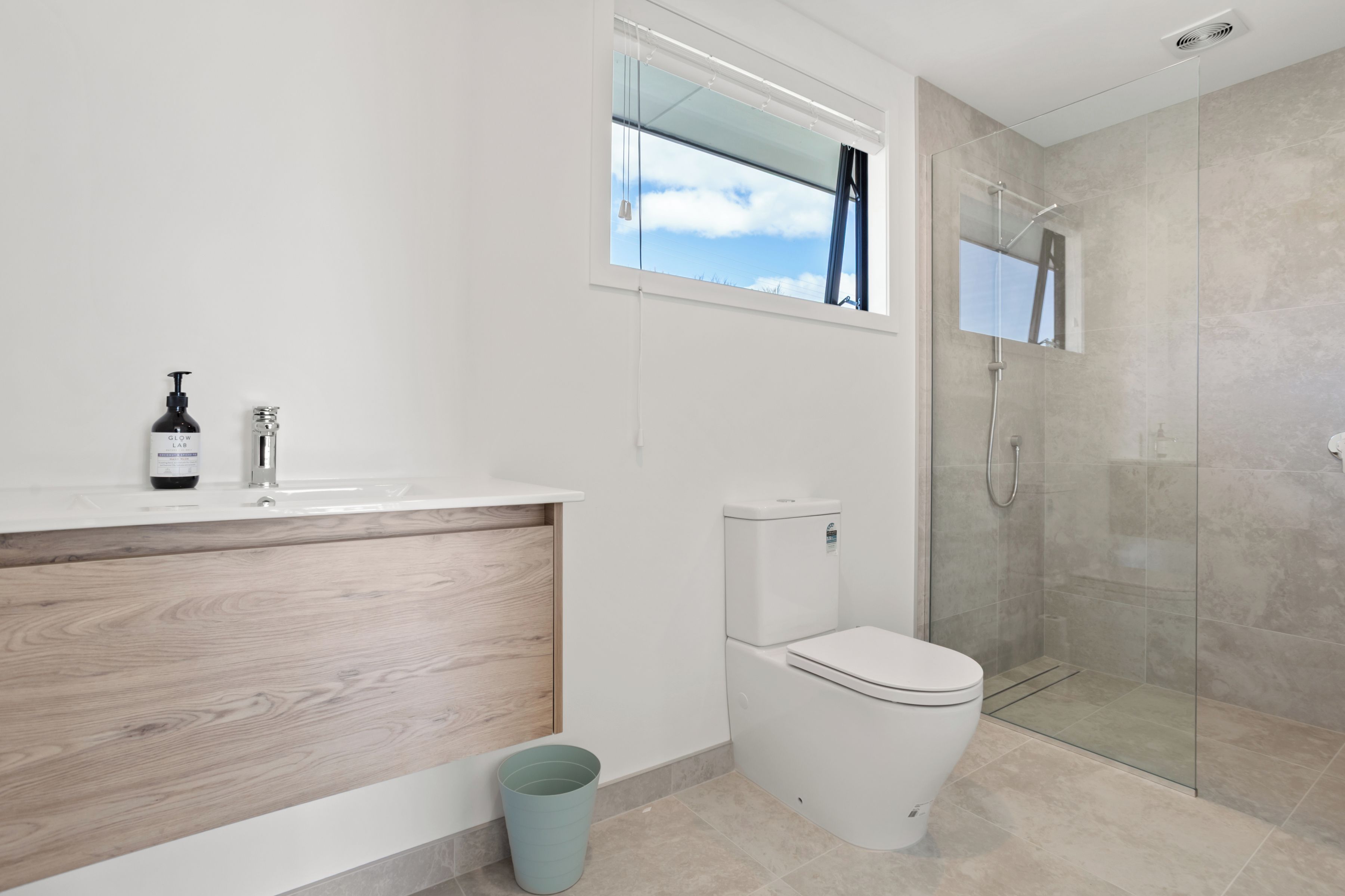 A modern bathroom with a wooden vanity, a toilet, a glass shower enclosure, and a window.