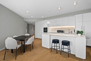 A modern kitchen with white cabinetry, a central island with bar stools, and a dining area with a dark table and white chairs. - Skyline 3-Bed Apartment Near South Melbourne (South Melbourne)