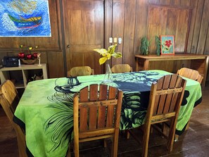 A dining area with a table covered in a tropical leaf pattern, wooden chairs, a vase with yellow flowers, and a painting on the wall.