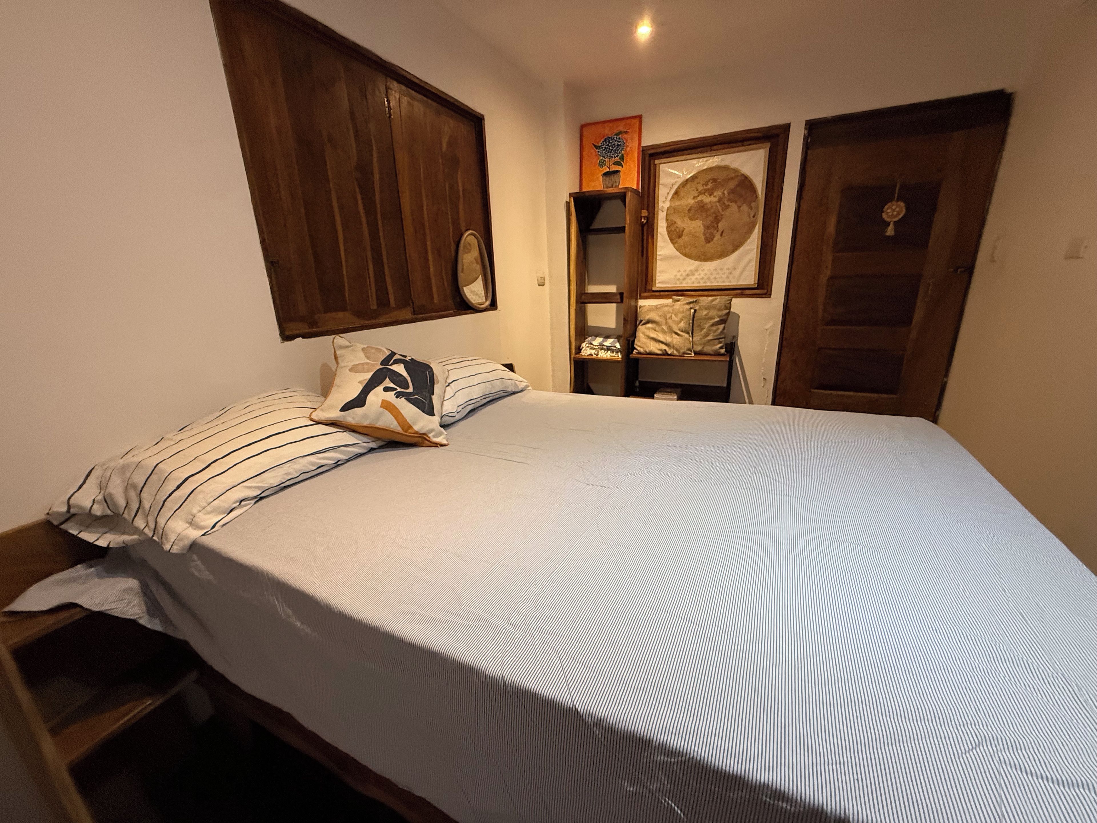 A bedroom with a bed, a wooden wardrobe, a framed picture on the wall, and a small shelf with a cushion.