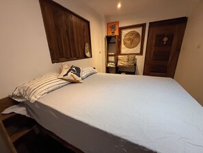 A bedroom with a bed, a wooden wardrobe, a framed picture on the wall, and a small shelf with a cushion.