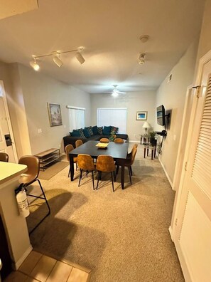 A dining area with a rectangular table and chairs, a ceiling fan, and a television mounted on the wall. - Full BR Resort near Disney/Pool, Jacuzzi, Splash, Lake, Club House/& Family Fun (Kissimmee)