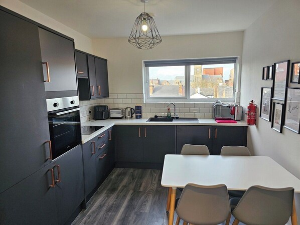 A modern kitchen with dark cabinetry, a dining area with a table and chairs, and a window with a view of buildings. - Market Quarter, town centre. free parking & wifi (Southport)