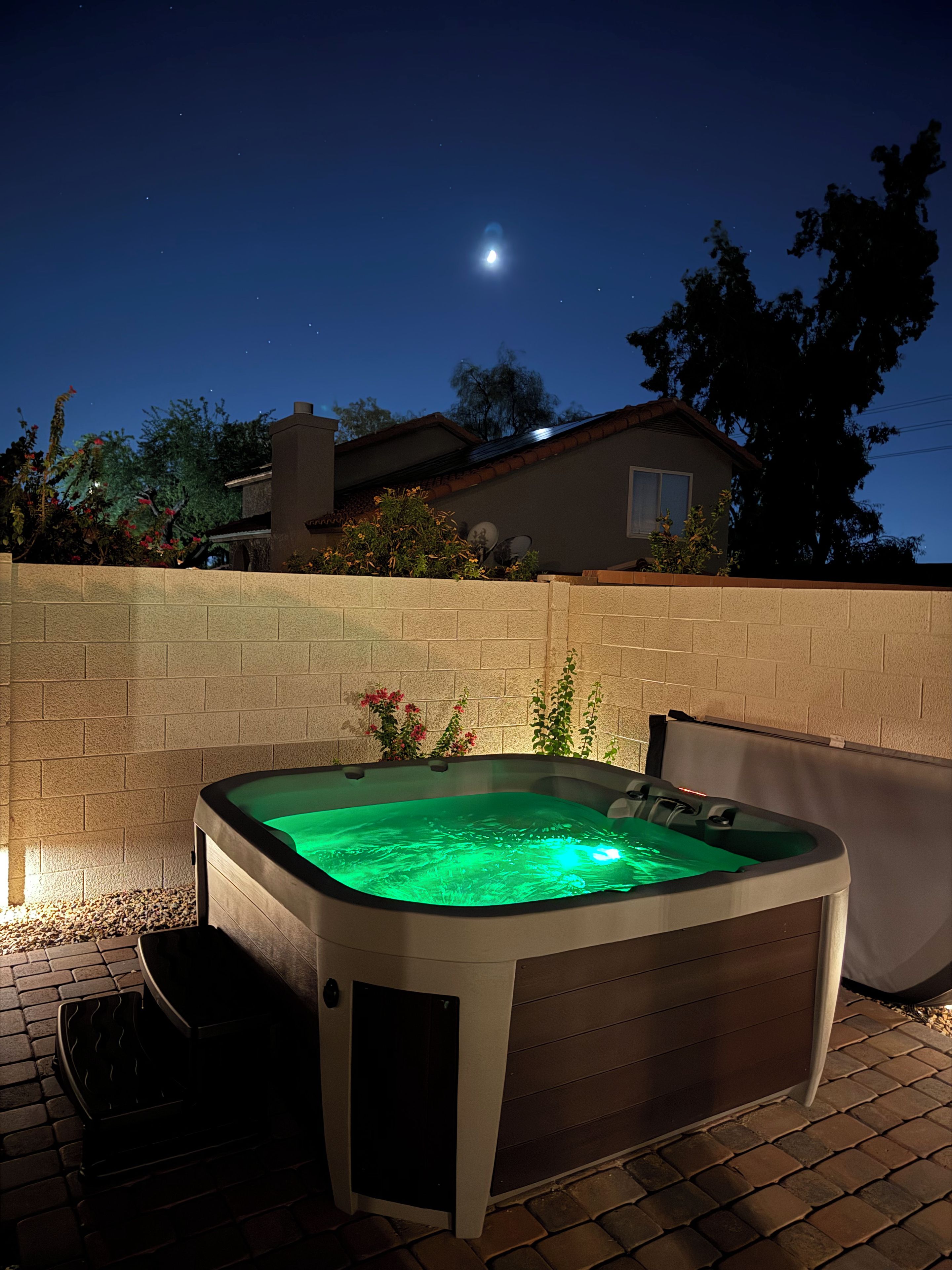 A hot tub with a green light, surrounded by a brick wall, with a house and trees in the background.