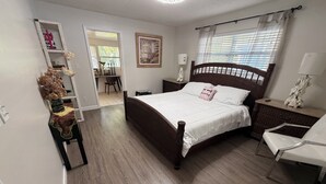 Quarto com cama de madeira, mesa de cabeceira, cadeira, criado-mudo, espelho e porta que dá para outro cômodo.