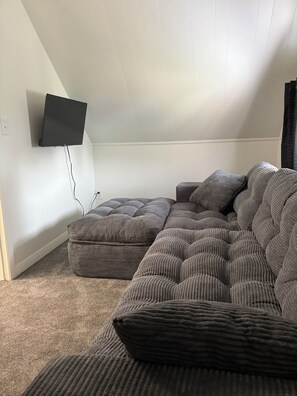 A room with a sectional sofa, a flat-screen TV mounted on the wall, and a carpeted floor.
