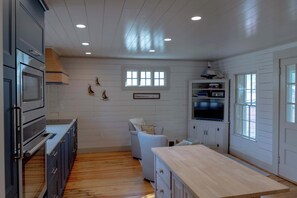 A kitchen with wooden cabinets, a built-in oven, a dining area with a wooden table, and a TV mounted above a cabinet. - Coastal Retreat Between Camden and Rockland (Rockport)