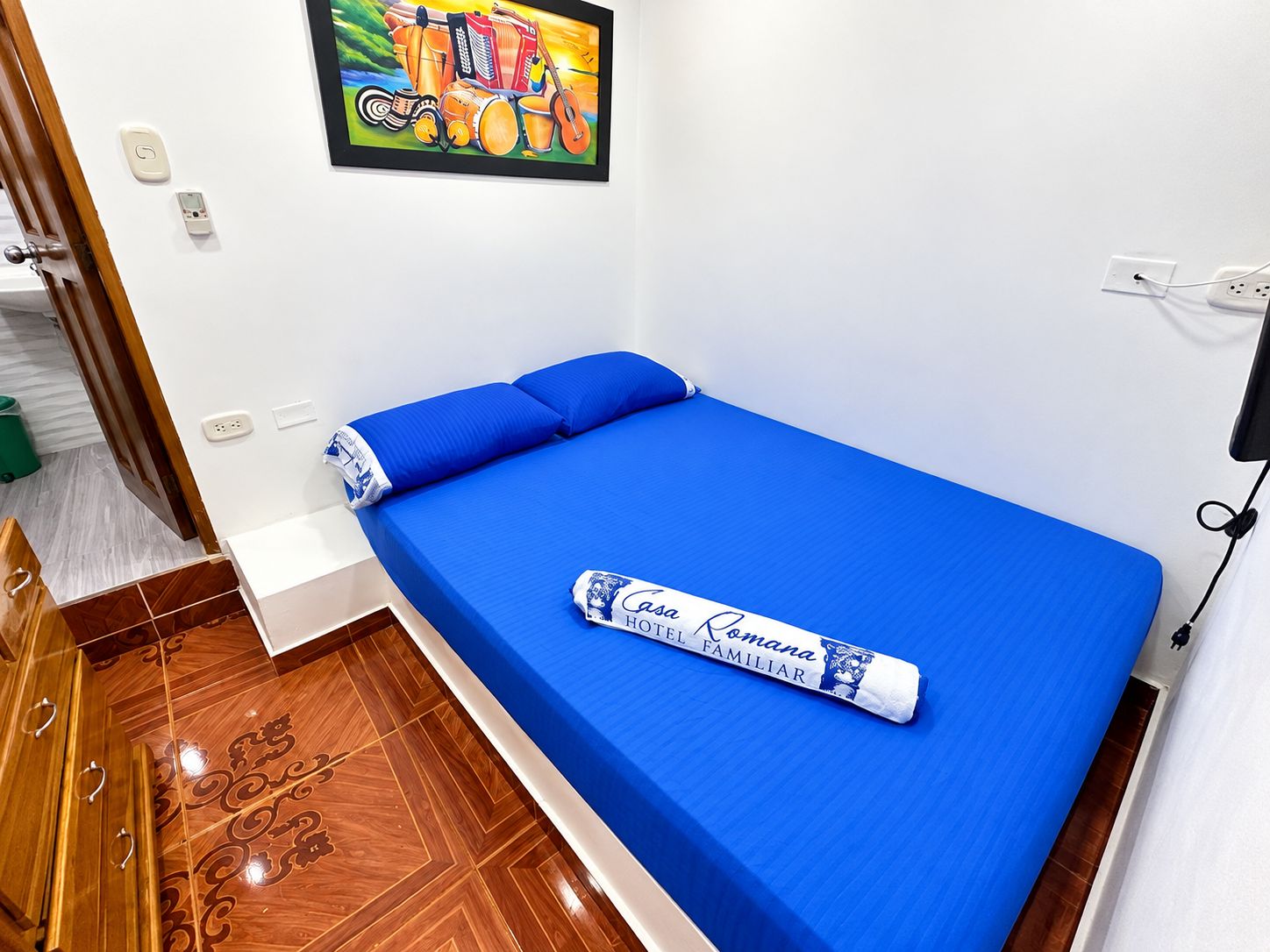 Uma cama de solteiro com colchão e almofadas azuis, uma toalha enrolada com texto, um quadro na parede e uma tomada.
