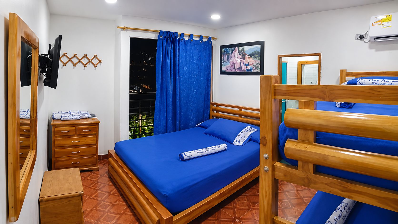 Una habitación con una cama, un aparador de madera, una mesita, un espejo, un ventanal con cortinas y un televisor montado en la pared.