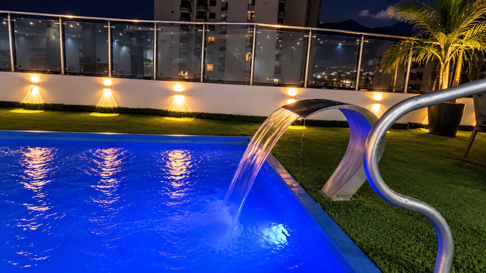 Una piscina con fuente, rodeada por una barandilla de vidrio e iluminada con luces ambientales.