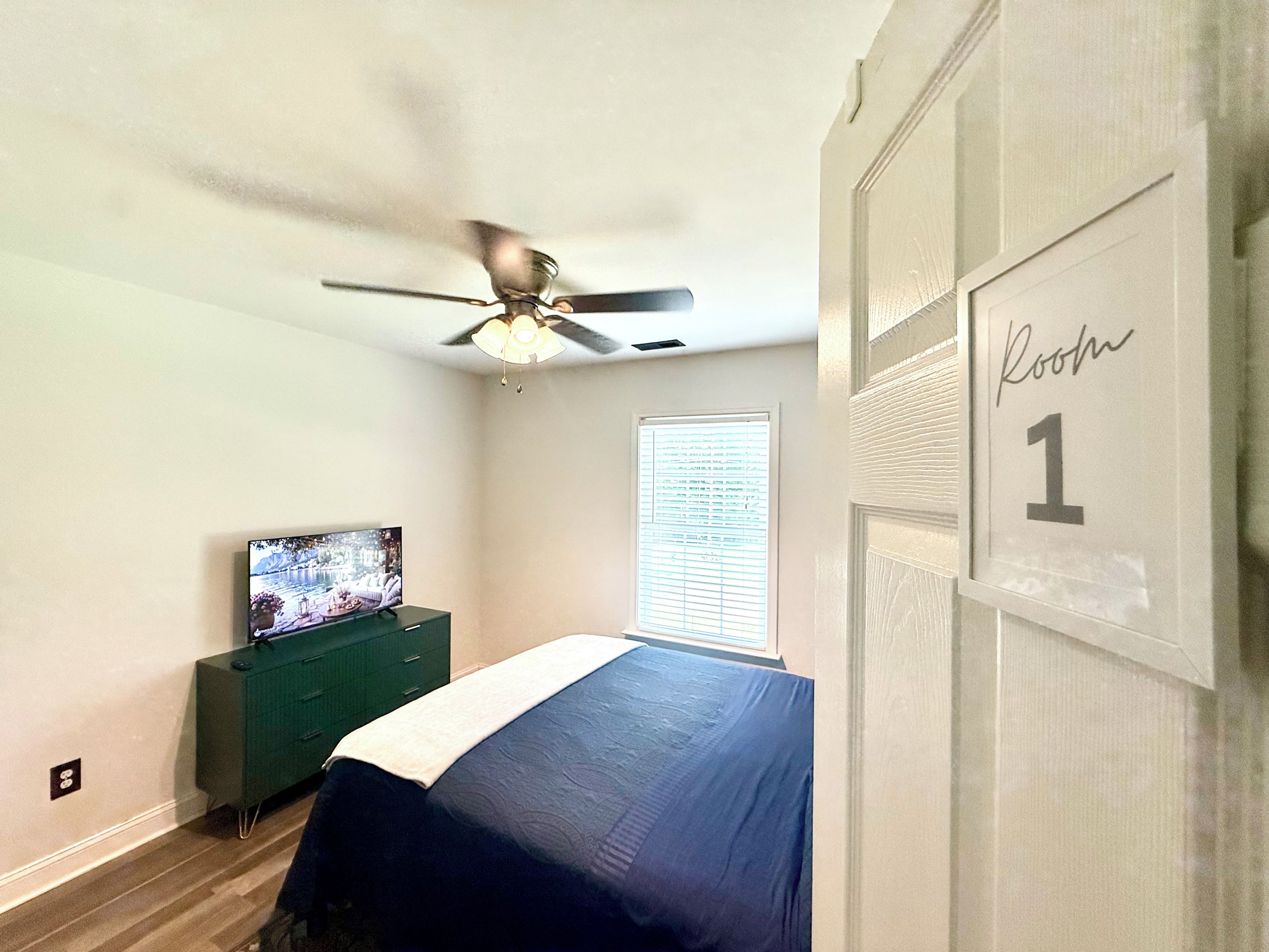 A bedroom with a bed, a television, a ceiling fan, and a door with a room number sign.