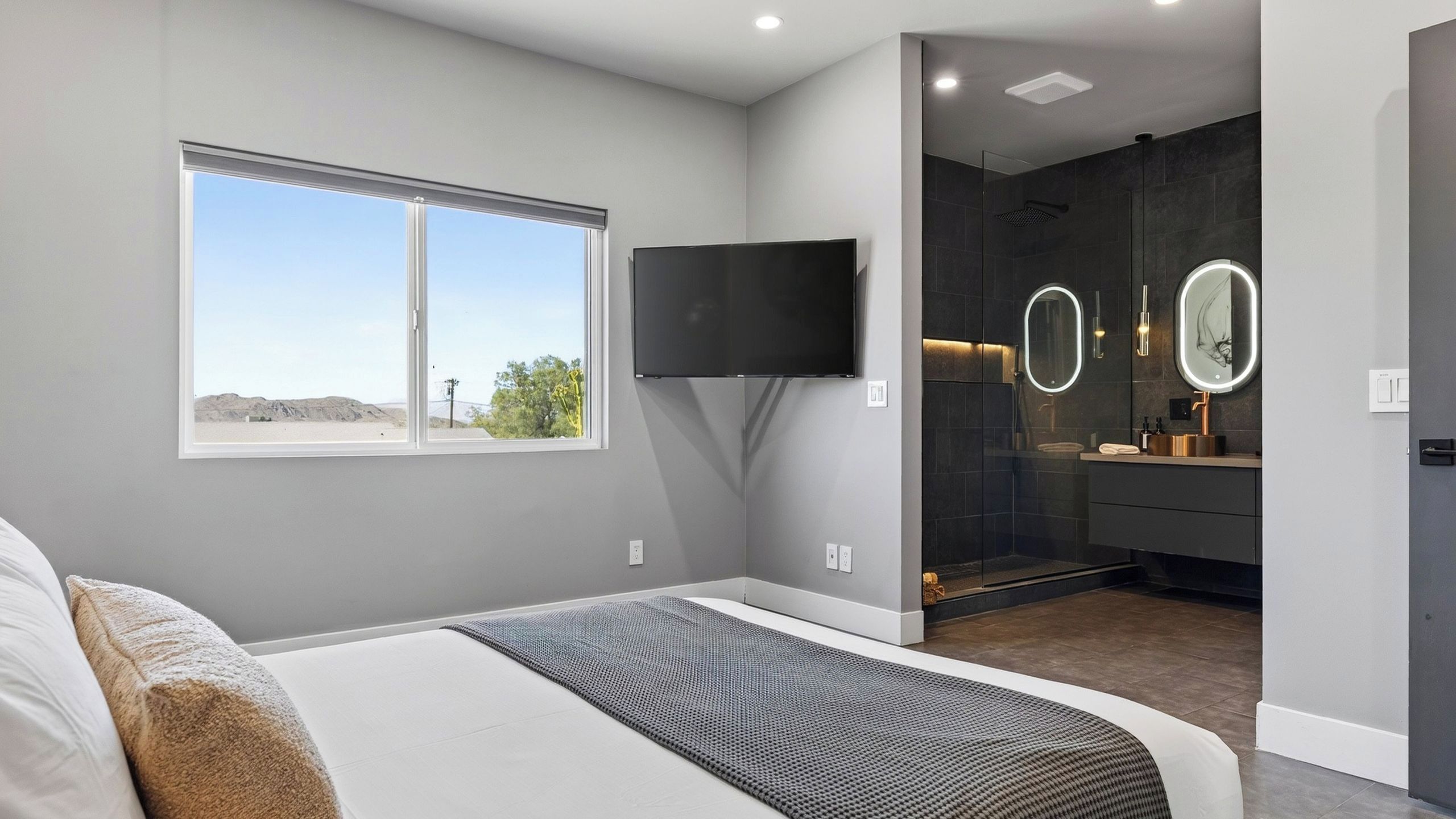 A modern bedroom with a large bed, a flat-screen TV mounted on the wall, and a window with a view of the outdoors.
