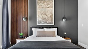 Ein modernes Schlafzimmer mit einem großen Bett, einer Wandpaneele aus Holz, einem gerahmten abstrakten Gemälde und zwei Pendelleuchten.