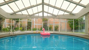 Una piscina interior con techo de cristal, un flotador de flamenco rosa y una pared de ladrillo al fondo.
