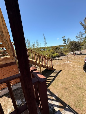 Eine Holzterrasse mit einer Treppe, die zu einem Gebäude führt, umgeben von einer trockenen, grasbewachsenen Fläche mit spärlicher Vegetation und blauem Himmel.
