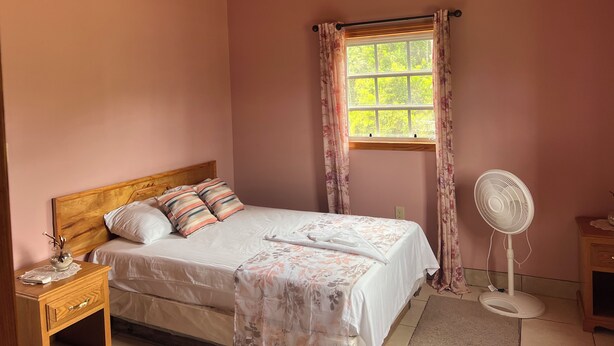 A bedroom with a bed, a bedside table, a nightstand, a window with curtains, and a standing fan. - perfect for family. Angler and nature guides (Queens highway)