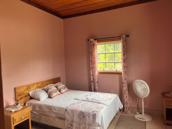 Ein Schlafzimmer mit einem Bett, einem Nachttisch, einem Fenster mit Vorhängen und einem Standventilator.