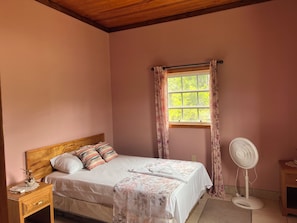 A bedroom with a bed, a bedside table, a nightstand, a window with curtains, and a standing fan.