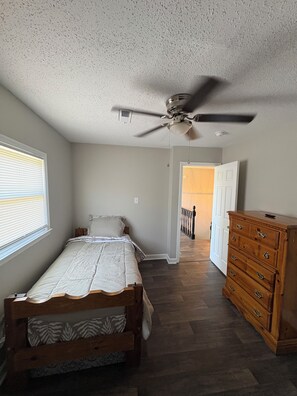 Ein Schlafzimmer mit Bett, Kommode, Deckenventilator und einem Fenster mit Jalousien.
