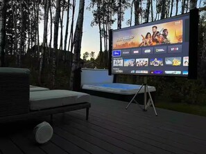 A large outdoor screen displaying a movie selection menu, with a wicker lounge chair and a speaker in the foreground.
