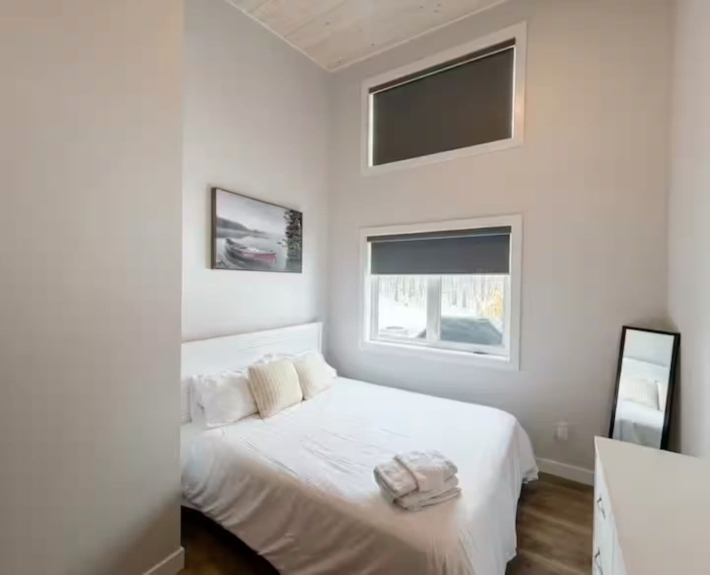 A bedroom with a bed, two windows, a painting, and a mirror.