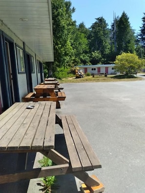 Un hilera de mesas de picnic de madera frente a un edificio, con cielo despejado y árboles al fondo.