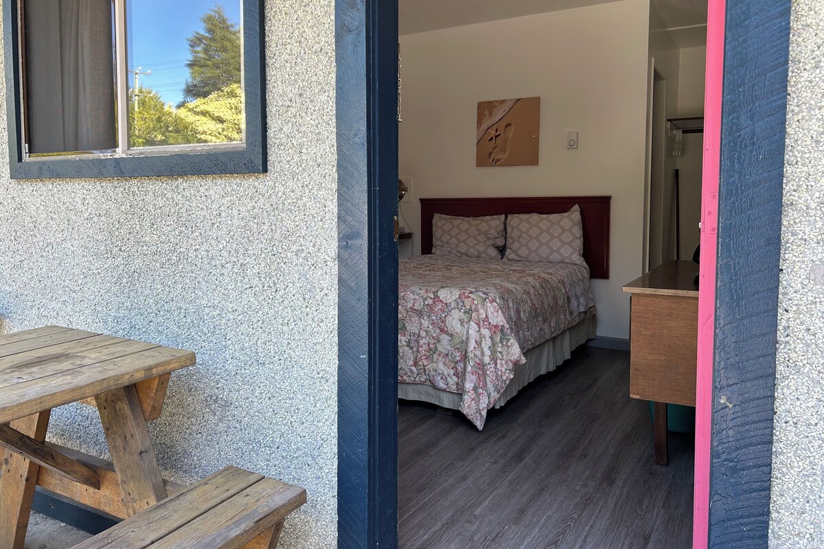 Un dormitorio con cama, una mesa de madera y un banco frente a un edificio con pared texturizada.