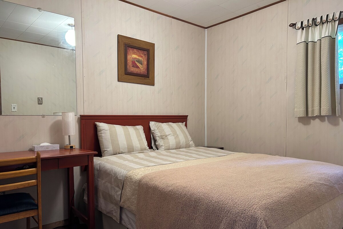 Una habitación de hotel con una cama, un escritorio, una silla, una mesita de noche, una lámpara, un cuadro enmarcado en la pared y una ventana con cortinas.