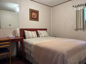 A hotel room with a bed, a desk, a chair, a bedside table, a lamp, a framed picture on the wall, and a window with curtains.