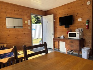 A wooden-paneled room with a door leading outside, a table with chairs, a microwave, a small cabinet, and a television mounted on the wall.