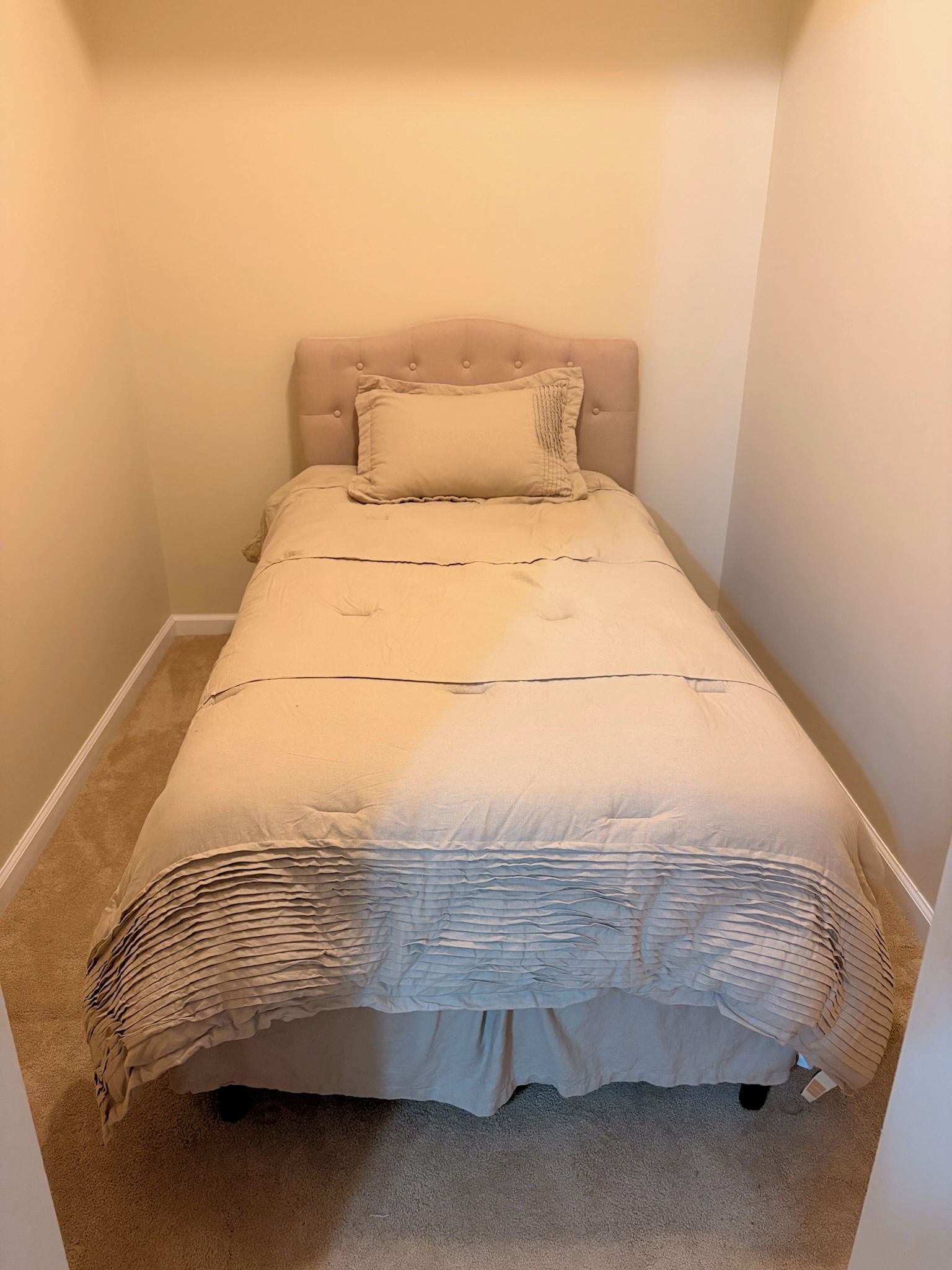 Et lille værelse med en enkelt seng, beige sengegavl og matchende pude. Sengen har et hvidt lagen og et vamset tæppe.