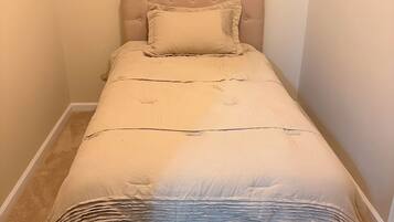 Een kleine kamer met een enkel bed, een beige hoofdbord en een passend kussen. Het bed heeft een wit laken en een gebreid plaid.