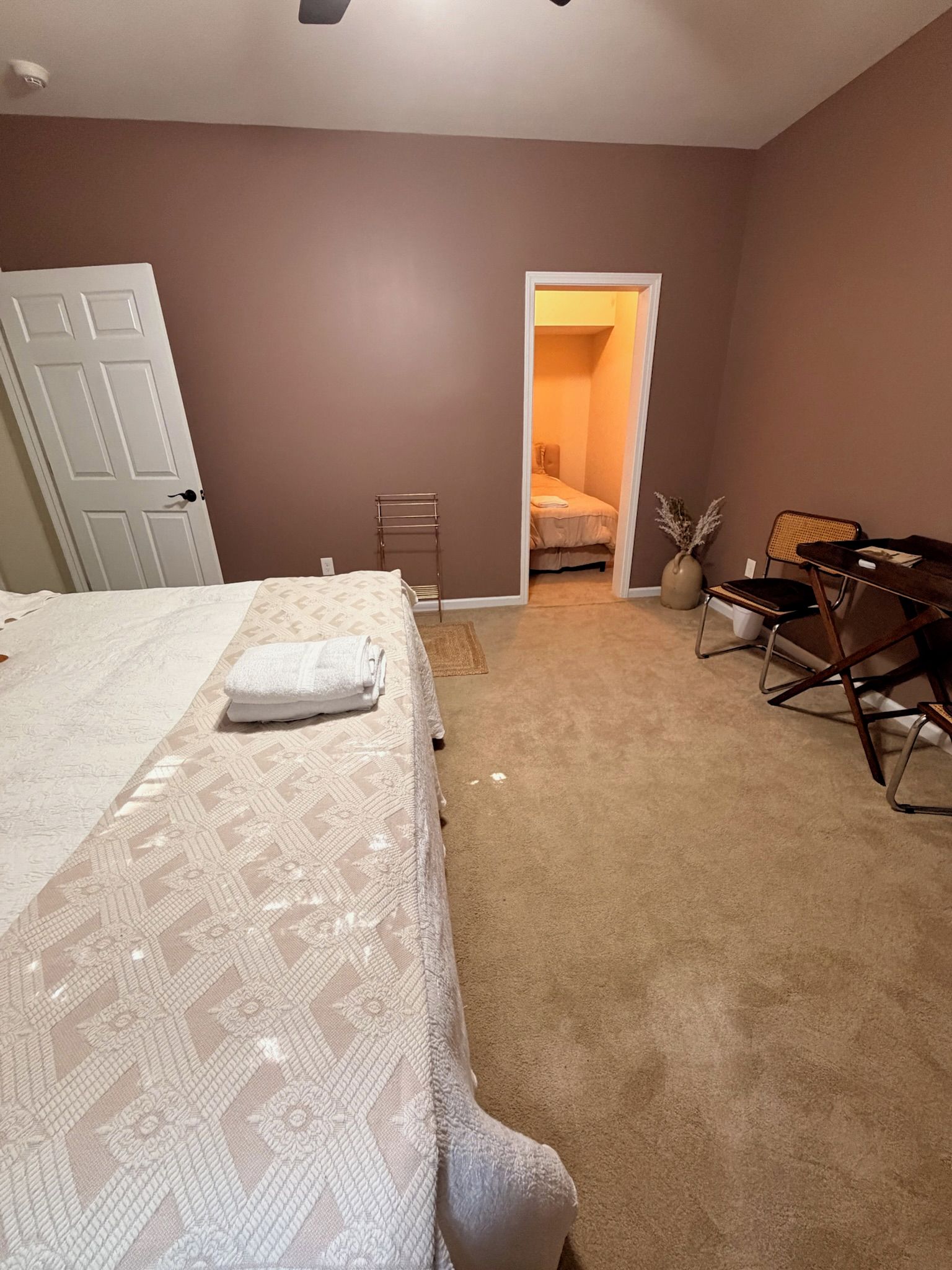 A bedroom with a bed, a desk, a chair, and a door leading to another room.