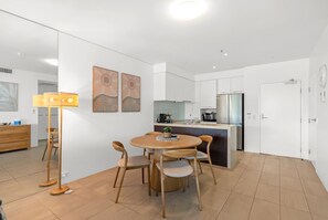 A modern kitchen with white cabinets, a central island, and a dining area with a round table and chairs. - Belle Escapes 3 Harbour Lights Marina View with Terrace (Cairns City)