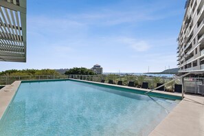 A rectangular swimming pool with a clear blue water, surrounded by a fence, and overlooking a distant view of buildings and a body of water. - Belle Escapes 3 Harbour Lights Marina View with Terrace (Cairns City)