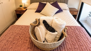 Ein Bett mit einer weißen und beige gestreiften Tagesdecke, einem violetten Kopfkissen und einem Korb mit Handtüchern und einem schwarzen Gegenstand auf einem Nachttisch aus Holz.