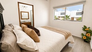 A bedroom with a bed, pillows, a bedside table, a window with a view of buildings, and a potted plant with sunflowers. - Villa Guacamaya Ocean View • Private Rooftop • Spacious 4-Bedroom Home (La Paz)