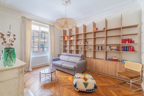 A well-lit living room with a sofa, a wooden bookshelf, a radiator, and a window with curtains. - Le Charme Chavannes - Welkeys (Paris)
