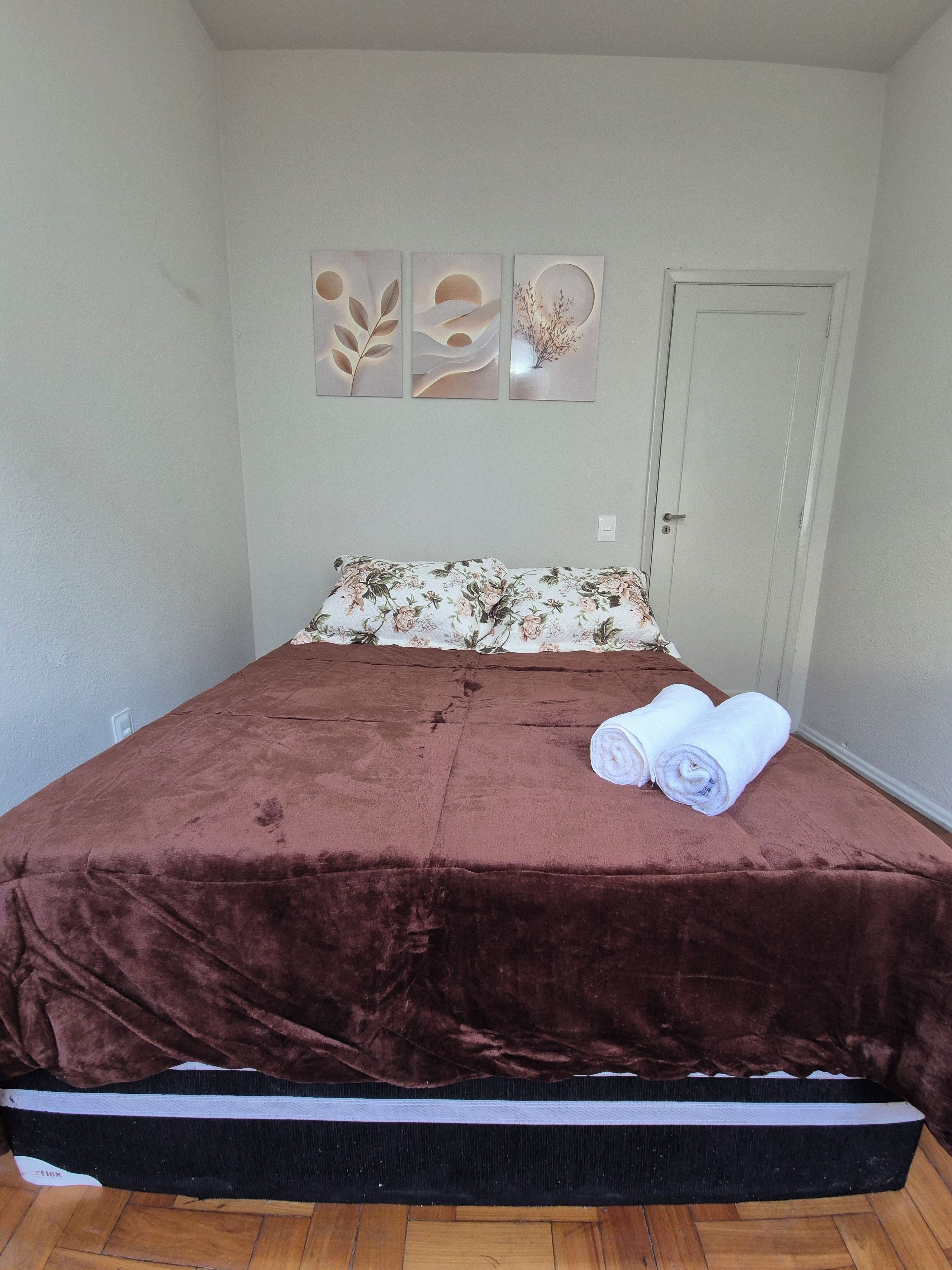 A single bed with a brown cover, a floral pillow, and two rolled towels on the bed. There are three wall art pieces above the bed.