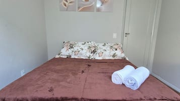 A single bed with a brown cover, a floral pillow, and two rolled towels on the bed. There are three wall art pieces above the bed.