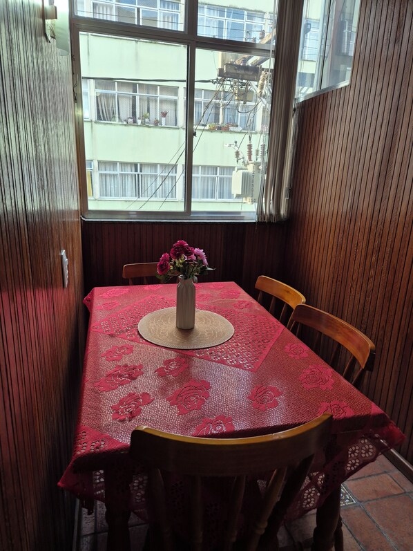 A dining area with a table set for two, a vase with flowers, and a view of buildings through a window.