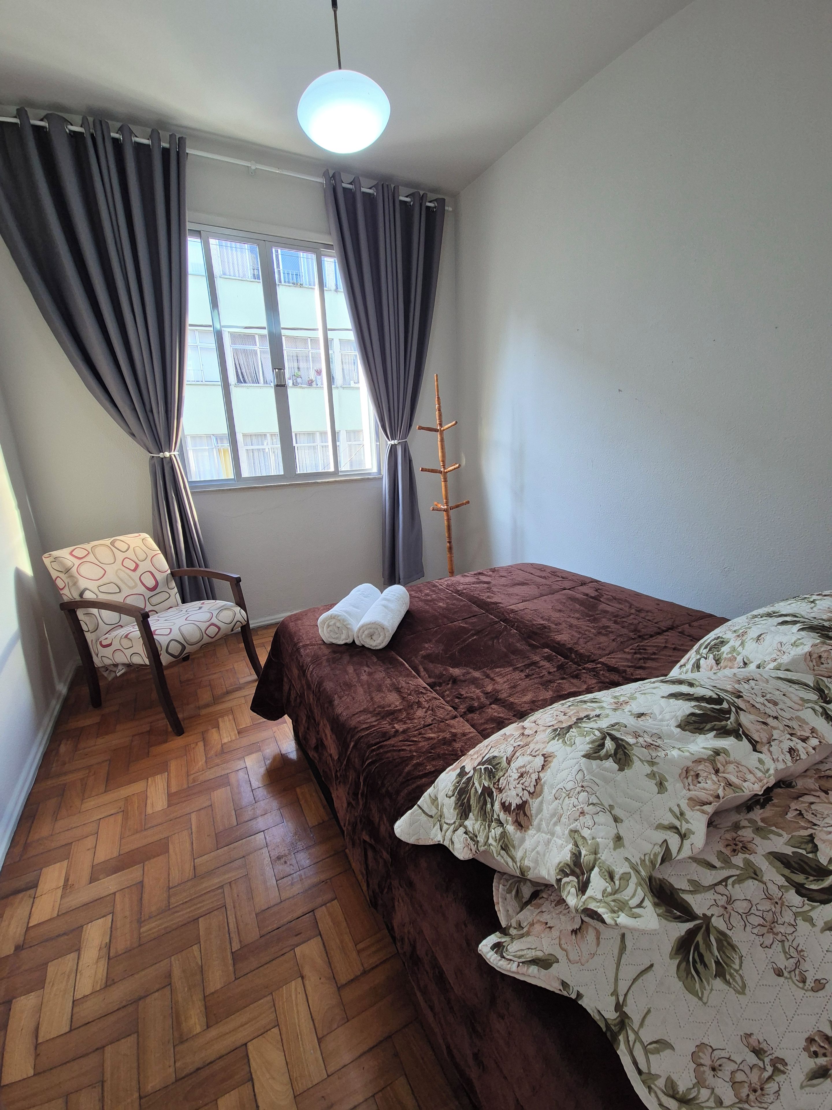 A bedroom with a bed, a chair, a window, and a hanging light fixture.