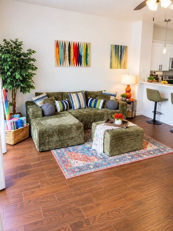 Ein Wohnzimmer mit einer Eckcouch, Poufs, einem gemusterten Teppich, einer Topfpflanze und Bildern an der Wand.