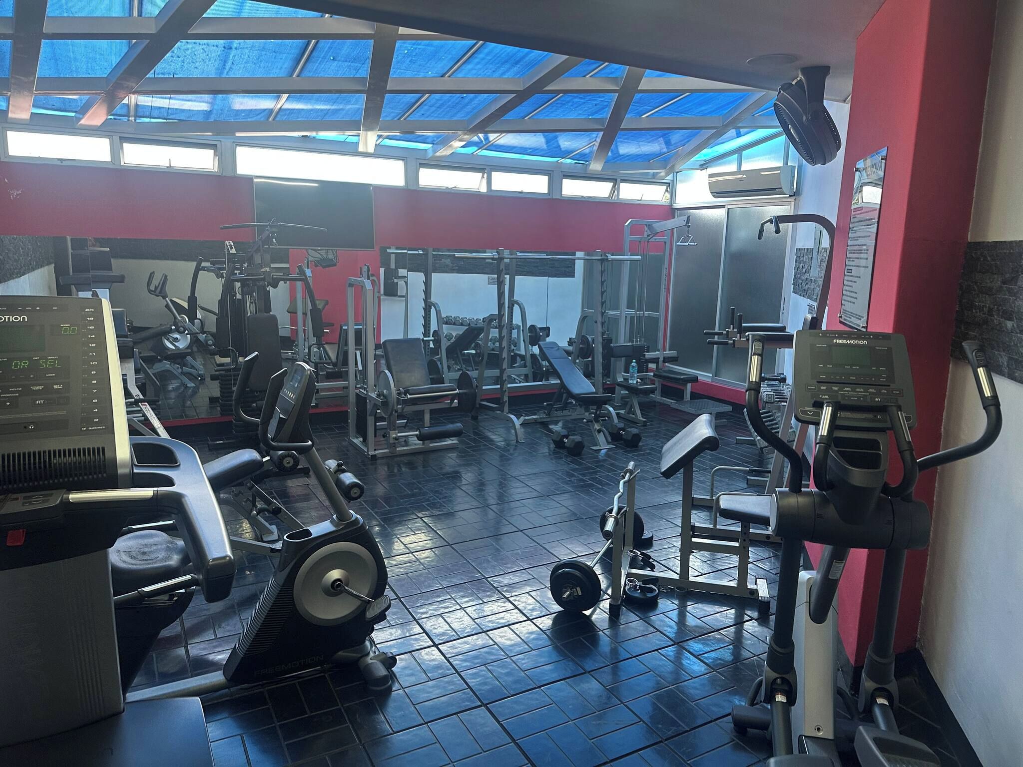 Ein Fitnessraum mit unterschiedlichen Trainingsgeräten, einer Decke mit Oberlichtern und einer roten Wand.