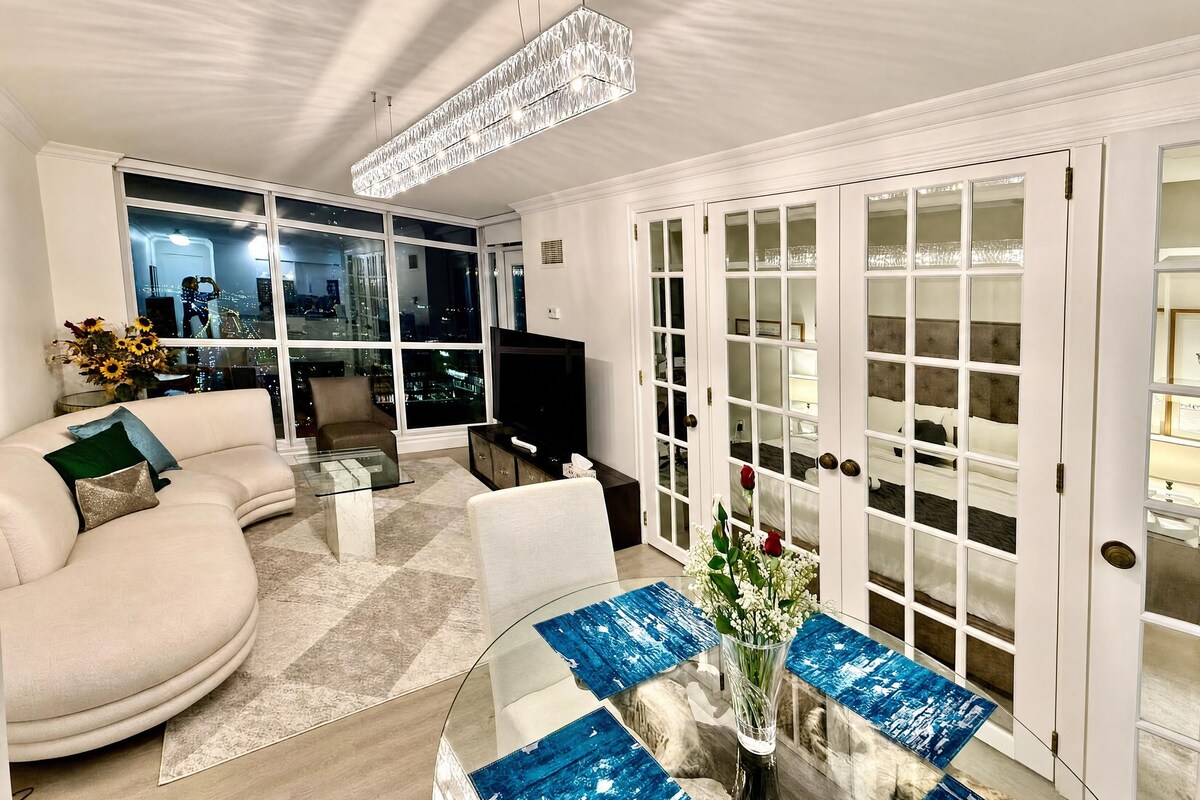 A modern living room with a sofa, coffee table, and a TV. There is a large window with a city view and a chandelier.