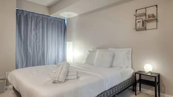 Una cama bien hecha con sábanas blancas, una mesita de noche con lámpara y una repisa en la pared con adornos.