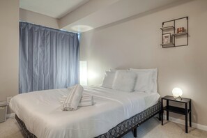 A neatly made bed with white linens, a bedside table with a lamp, and a wall-mounted shelf with decorative items. - South Core Suites (Toronto)